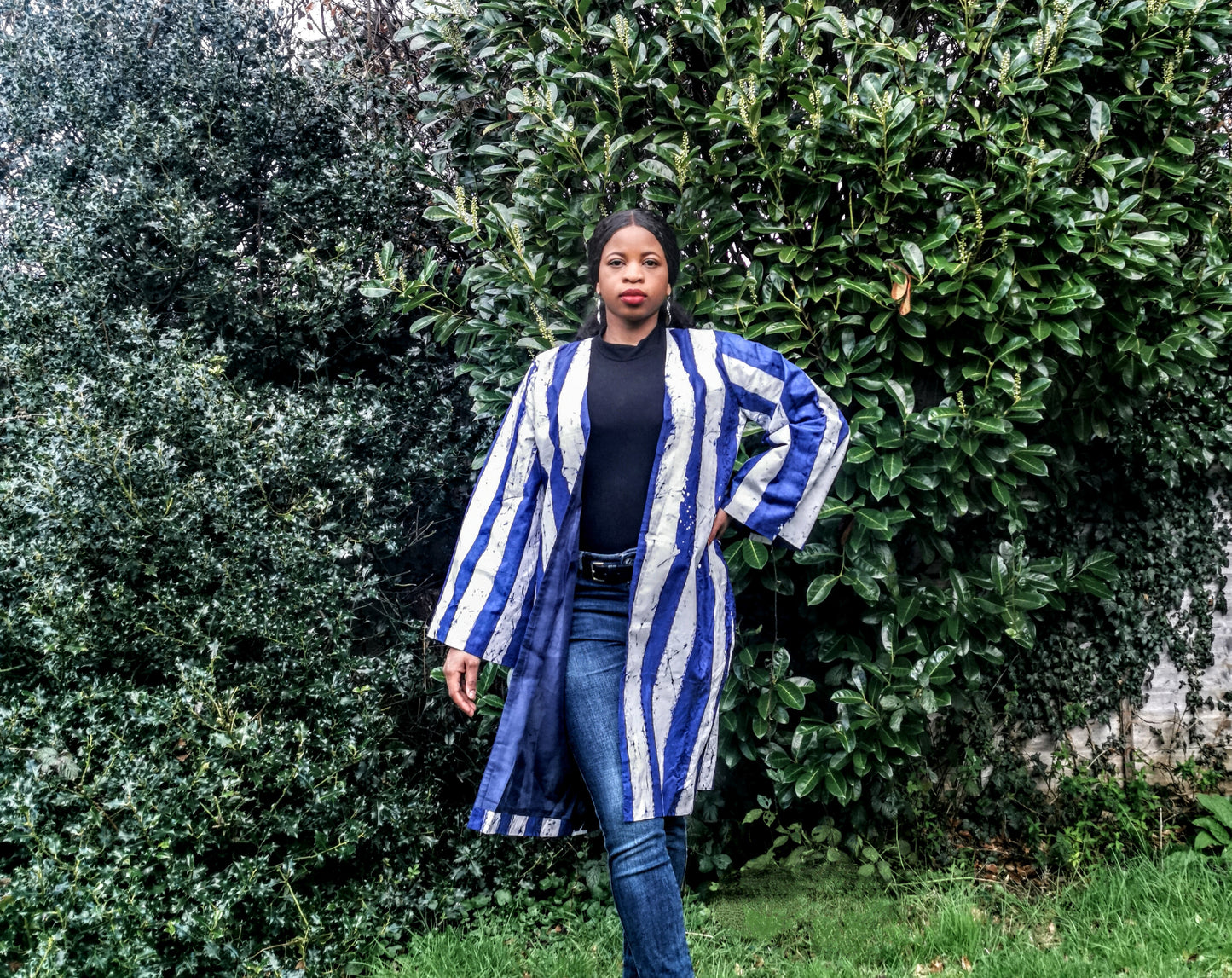 Woman wearing a blue and white striped kimono standing in front of green bushes.